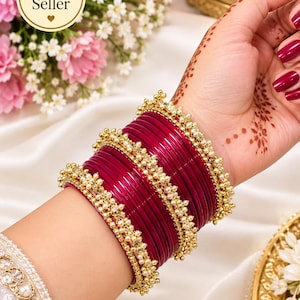 May include: A set of deep magenta bangles featuring a smooth, glossy finish. The stack includes several plain, thin bangles interspersed with two wider, gold-toned bangles adorned with intricate clusters of small, white, pearl-like beads.
