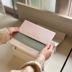 May include: A set of rectangular stationery cards in pale pink, sage green, and cream, stacked inside a matching box. Each card features the gold-embossed text "BEST WISHES" followed by the names "Kallana & Sayanak Sangal" in a cursive script.