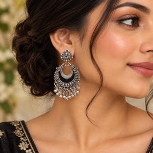 May include: A silver-toned dangle earring featuring a circular stud top and a large crescent-shaped drop. The crescent is accented with a smooth black inlay and intricate metalwork, finished with a row of small dangling silver beads at the base.