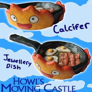 May include: A black frying pan shaped jewellery dish featuring a sculpted orange and yellow fire character with white eyes and a red mouth. Inside the pan are two fried eggs and strips of bacon. The text reads Calcifer, Jewellery Dish, Howl's Moving Castle.