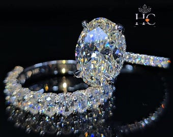 Anel de noivado em ouro branco com moissanita oval de 3 quilates, aro cravejado de diamantes, aliança de casamento oval alongada combinando e anel de noivado com halo oculto.
