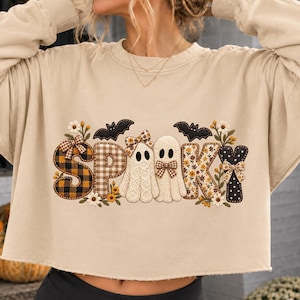 May include: A beige long-sleeve cropped sweatshirt featuring a graphic print that spells SPOOKY. The letters are decorated with plaid, floral, and polka dot patterns, small bows, and white flowers. Two white ghost figures and black bats are included.