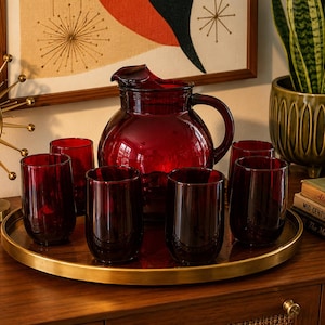 May include: A deep red glass pitcher and five matching tumblers arranged on a circular gold-toned metal tray. The pitcher features a rounded body with a curved handle and a pouring spout, while the tumblers have a simple, cylindrical shape.