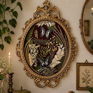 May include: An oval wall frame with an ornate gold finish containing a dark red velvet backing. The display features two black and white butterflies surrounded by an arrangement of dried green ferns, white flowers, and tall grasses.
