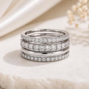 May include: A stack of four silver-toned rings. The top and bottom rings feature a pavé setting with small round stones. The second ring from the top has a channel setting with square-cut stones, and the third ring is a plain, polished band.