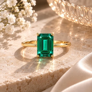 May include: A gold ring featuring a large, rectangular emerald-cut green gemstone set in a four-prong setting. The band is thin and polished gold, showcasing a vibrant, deep green center stone with sharp, clean facets that reflect light.