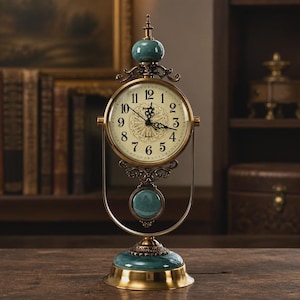 Puede incluir: Un reloj de mesa con esfera redonda color crema, números arábigos negros y manecillas ornamentadas. El reloj está encajado en un marco de metal dorado con detalles de piedra color verde azulado en la parte superior, central y base.