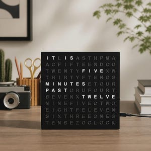 Peut inclure: Une horloge à mots carrée noire avec une grille de lettres. Les mots IT IS, MINUTES, PAST, FIVE et TWELVE sont éclairés en blanc vif sur le fond sombre, indiquant l'heure actuelle. Un câble d'alimentation noir sort sur le côté droit.