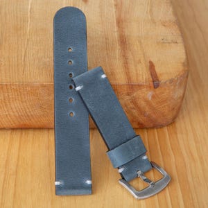 May include: A dark gray leather watch strap featuring a smooth matte finish, white contrast stitching at the ends, and a silver-toned metal buckle. The strap consists of two pieces, one with six circular adjustment holes and one with a metal pin.
