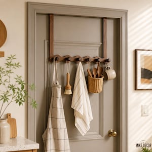 May include: A dark brown wooden over-the-door hook rack with six pegs. Hanging from the hooks are a beige grid-patterned apron, a wooden scrub brush, a white striped kitchen towel, a woven wicker basket with wooden spoons, and a ceramic mug.