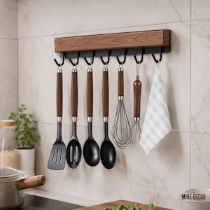 May include: A wall-mounted kitchen utensil rack featuring a dark wood grain horizontal bar with seven black metal hooks. Hanging from the hooks are a slotted turner, a slotted spoon, two solid spoons, two wire whisks, and a white striped towel.