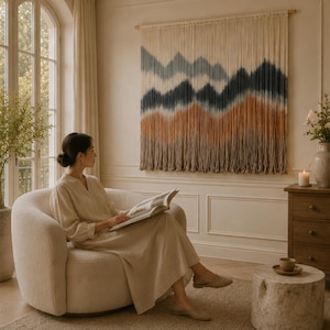 Op de afbeelding: Een grote rechthoekige macramé wandhanger met een berglandschapontwerp. Het textielkunstwerk toont horizontale kleurlagen in tinten crème, gedempt blauw, donker antraciet en terracotta, opgehangen aan een houten roede.