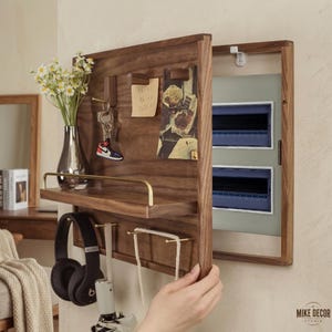 Op de afbeelding: Een wandkast van donker hout met een scharnierende deur die een meterkast verbergt. De buitenkant is voorzien van een plank met een messing reling, diverse haken voor sleutels en koptelefoons, en een oppervlak voor notities en foto's.