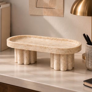 May include: An oval-shaped beige stone tray with a porous, textured surface. The tray features a flat, elongated top supported by two thick, cylindrical pedestals with a vertical fluted or scalloped design, crafted from a natural stone material.