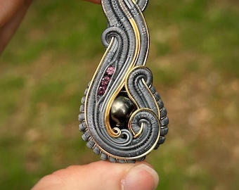 Può includere: Un ciondolo fatto a mano con un design a spirale, con filigrana grigia e dorata. Il ciondolo incorpora tre piccole pietre preziose rosa e una pietra sferica più grande e scura. L'intricato avvolgimento del filo crea un pezzo artistico unico.