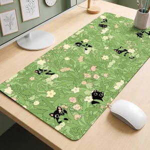 May include: A rectangular desk mat featuring a repeating pattern of black cats peeking out from a dense field of light green foliage and small white and pale pink flowers. The mat has a smooth surface and rounded corners, covering a wooden desk surface.