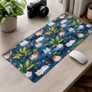 May include: A rectangular desk mat featuring a dark blue background with a repeating pattern of small cartoon cats swimming among green tropical leaves and tiny red flowers. The cats are depicted in various colours including orange, white, and grey.