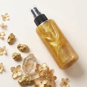 Könnte beinhalten: Eine durchsichtige Kunststoffsprühflasche gefüllt mit einer schimmernden goldenen Flüssigkeit und einem schwarzen Pumpkopf. Die Flasche ist umgeben von mehreren kleinen, strukturierten goldfarbenen Steinen, einem klaren Quarzkristall und Blütenblättern.