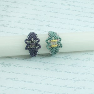 May include: Two handmade macrame rings with beaded accents. The ring on the left is purple and the ring on the right is green.