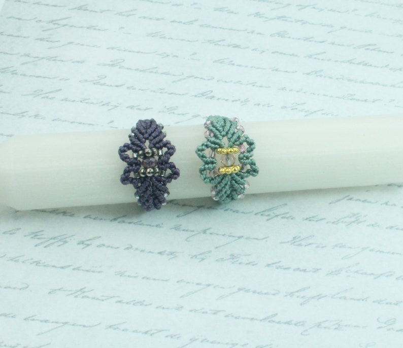 May include: Two handmade macrame rings with beaded accents. The ring on the left is purple and the ring on the right is green.