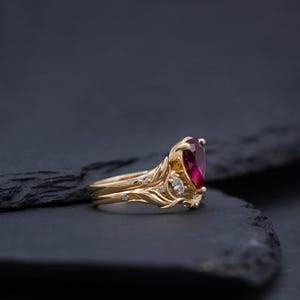 Puede incluir: Un anillo de oro con una piedra preciosa roja en forma de pera como pieza central, acentuada por piedras claras más pequeñas. La banda presenta detalles metálicos intrincados y elevados en forma de hoja que envuelven el engaste central.