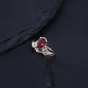 Può includere: Un anello in argento con una pietra preziosa rossa centrale a forma di pera, affiancata da due pietre chiare più piccole. La fascia in metallo presenta dettagli intricati, organici e simili a viticci che si estendono dalla montatura.