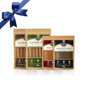 Pode incluir: Quatro bolsas verticais de especiarias e paus de canela com um laço azul no topo. Os rótulos apresentam o texto Ceylon, Ceylon-Z e Schwarzer Pfeffer, contendo cada bolsa 50g de produto. A embalagem é de papel castanho com janelas transparentes.