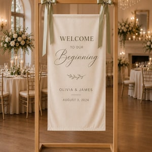 May include: A rectangular cream-colored fabric sign suspended from a light wood frame by two sage green ribbons. The sign features the text "WELCOME TO OUR Beginning", a small leaf graphic, "OLIVIA & JAMES", and the date "AUGUST 3, 2024" in dark lettering.