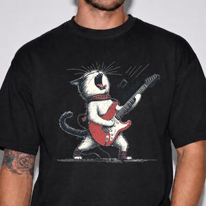 Puede incluir: Una camiseta negra de manga corta con una ilustración gráfica de un gato blanco con bufanda roja tocando una guitarra eléctrica roja. El gato tiene la boca abierta como si estuviera cantando, con notas musicales en el aire sobre el instrumento.