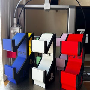 Puede incluir: Una colección de soportes verticales para mandos de juego en azul, blanco y rojo, cada uno con un marco negro, el logotipo de PS5 en la parte frontal y el símbolo de PlayStation en la base. Estos soportes mantienen los mandos seguros.