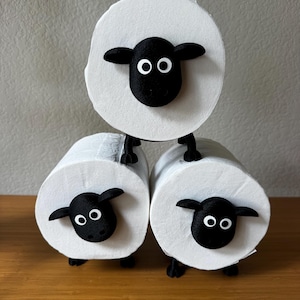 May include: Three white toilet paper rolls decorated to resemble sheep. Each roll features a black 3D-printed sheep head with white eyes and small black legs attached to the bottom, creating a stacked arrangement of three individual sheep figures.