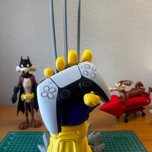 May include: A 3D-printed display stand shaped like a yellow-gloved hand with three long silver claws, holding a white and black wireless game controller. The base is yellow with brown accents and gray rock-like details, featuring a cartoon character figure.