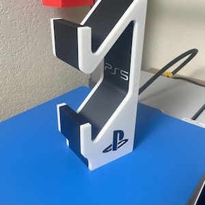 May include: A white and black vertical gaming controller stand featuring the PS5 logo printed on the side and the PlayStation symbol at the base. The stand has a tiered design with two cradles for holding a controller, constructed with a matte finish.