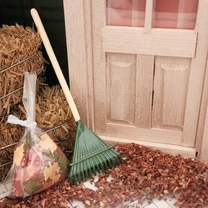 May include: A miniature garden rake with a long, light-colored wooden handle and a dark green plastic head, positioned next to a small, clear plastic bag filled with assorted autumn-colored leaf cutouts and a light-colored wooden door frame.