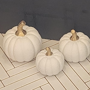 May include: Three decorative pumpkins in varying sizes, each featuring a matte white finish and a metallic gold stem. The pumpkins have a rounded, ribbed shape and are arranged in a cluster, showcasing a consistent design across all three pieces.