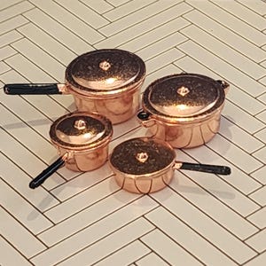May include: A set of four miniature copper-colored pots and pans with lids. Two pieces feature long black handles, while the other two have small side handles. The metallic surfaces have a reflective finish and the lids include small central knobs.