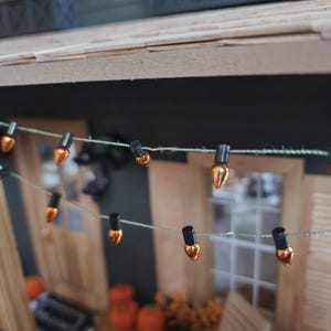 May include: A string of miniature decorative lights featuring small, metallic orange, teardrop-shaped bulbs attached to black bases. The bulbs are connected by a thin, light green string, creating a festive garland effect for a miniature display.