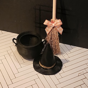 May include: A miniature black cauldron with two side handles, a small black pointed hat with a gold band, and a miniature broomstick featuring a light wooden handle and a tan straw brush tied with an orange and white checkered fabric bow.