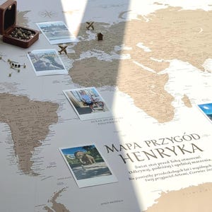 Può includere: Una grande mappa del mondo beige con continenti marroni, una rosa dei venti, una piccola scatola di legno contenente spilli, diversi piccoli segnaposto a forma di aereo in legno e varie foto quadrate. Il testo riporta MAPA PRZYGÓD HENRYKA.
