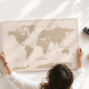 Può includere: Un poster rettangolare raffigurante una mappa del mondo con sfondo beige e continenti color sabbia, recante la scritta "MAP OF THE WORLD" al centro in basso. La mappa include una rosa dei venti ed è mostrata accanto a un piccolo mappamondo.