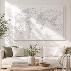 Könnte beinhalten: Ein großes rechteckiges Wandbild mit einer detaillierten schwarz-weißen Weltkarte und einer Windrose in der unteren linken Ecke, das in einem dünnen weißen Rahmen an einer Wand über einem weißen Sofa mit dekorativen Kissen befestigt ist.