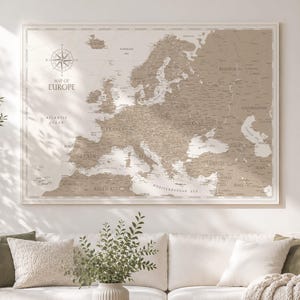 Könnte beinhalten: Eine rechteckige Wandkarte von Europa in neutralen Beige- und Weißtönen. Die Karte zeigt geografische Grenzen, Ländernamen und größere Gewässer. In der oberen linken Ecke befindet sich eine Windrose mit dem Schriftzug MAP OF EUROPE.