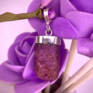 May include: A raw, deep red ruby gemstone pendant set in a polished silver cap with a circular bail. The stone has a rough, textured surface and a rectangular shape. The pendant is suspended from a thin, light brown stem among purple flowers.