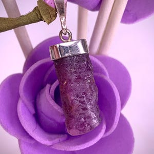 Puede incluir: Un colgante de piedra preciosa en bruto de color rojo púrpura intenso, engastado en una tapa de metal plateado pulido con una anilla a juego. La piedra tiene una superficie rugosa y texturizada y forma cilíndrica, suspendida de un aro metálico.