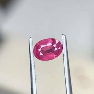 Puede incluir: Una gema rosa de corte ovalado sostenida por unas pinzas de metal plateado. La piedra presenta una superficie facetada que refleja la luz, mostrando un tono magenta profundo y saturado con claridad interna y un acabado brillante.
