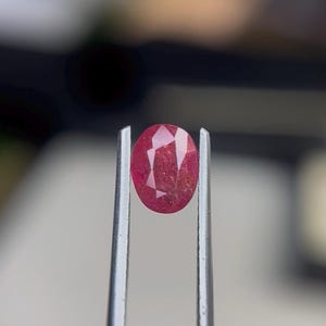Puede incluir: Una piedra preciosa de color rojo rosado vibrante, tallada en forma ovalada con múltiples facetas que reflejan la luz. La piedra se sostiene de forma segura entre las dos puntas finas de metal plateado de unas pinzas, mostrando su claridad.