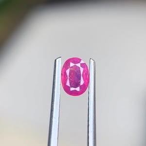 Puede incluir: Una gema rosa de corte ovalado sostenida firmemente por unas pinzas de metal plateado. La piedra presenta una superficie facetada que refleja la luz, mostrando un tono magenta intenso con claridad interna y un patrón geométrico distintivo.