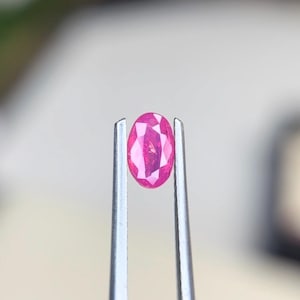 Puede incluir: Una gema rosa de corte ovalado con una superficie facetada que refleja la luz, sostenida firmemente entre las dos puntas metálicas de unas pinzas de tono plateado. La piedra muestra un tono magenta profundo y saturado con claridad.