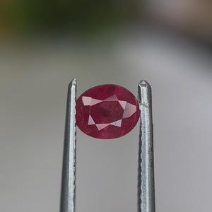 Puede incluir: Una piedra preciosa de rubí de forma ovalada y facetada, de color rojo intenso, sostenida entre las puntas metálicas dentadas de unas pinzas. La gema muestra múltiples facetas triangulares y trapezoidales en toda su superficie.