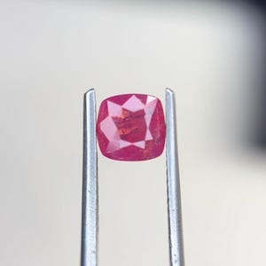 Puede incluir: Una piedra preciosa de color rosa vibrante con corte cojín, sostenida por unas pinzas de metal plateado. La piedra presenta una superficie facetada que refleja la luz, mostrando un tono magenta intenso con claridad interna y patrones geométricos.